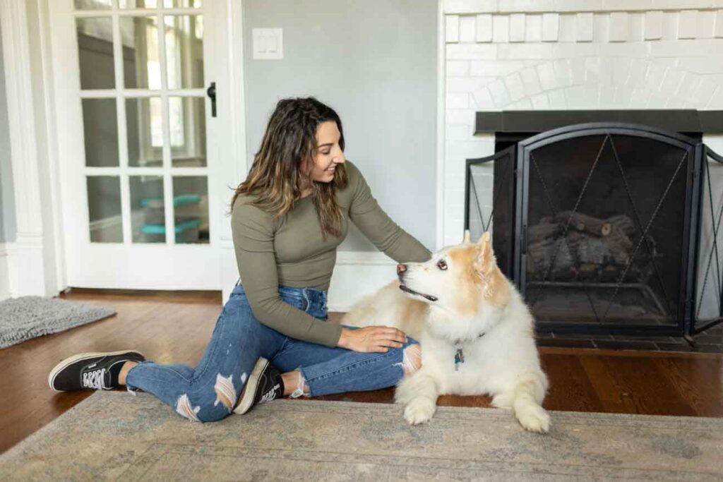 Woman and her husky look at each other while sitting in front of a white fireplace during an end-of-life photo session