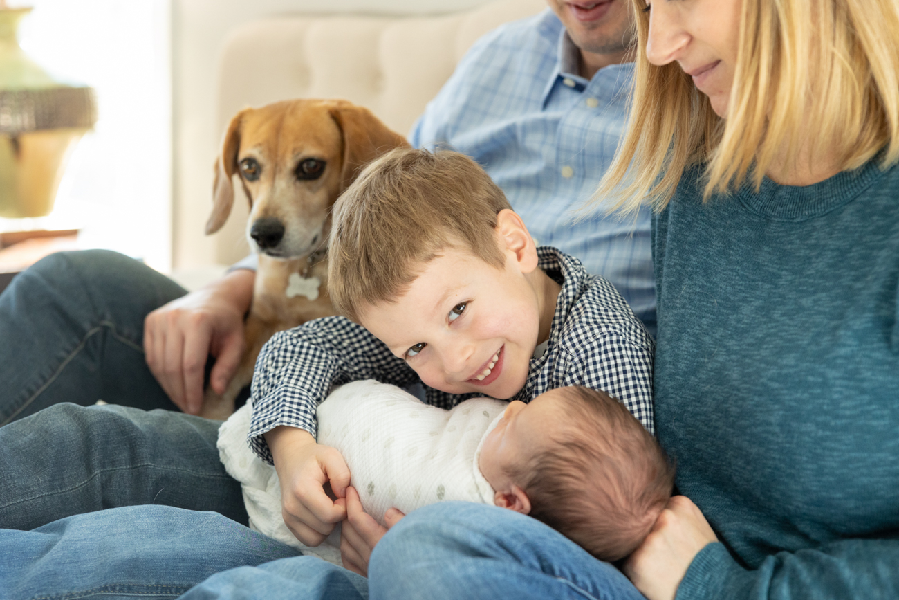Toddler boy smiles at the camera while holding newborn baby with his parents and dog looking on