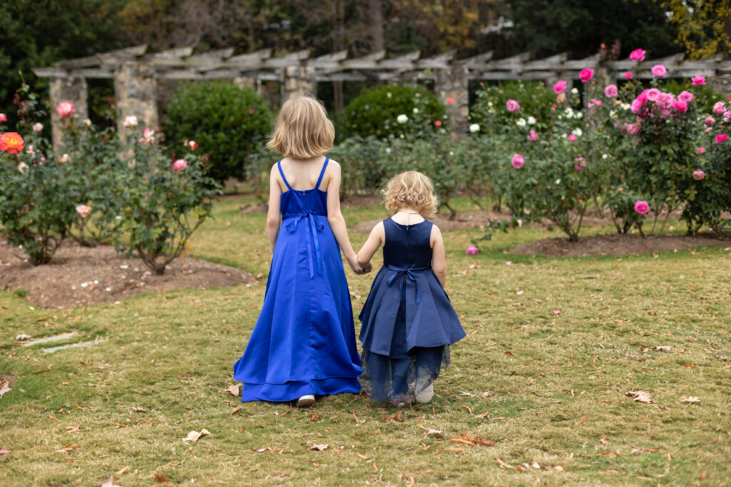 Two blond girls walk in a rose garden with blue formal dresses. Photo taken as the girls walk away from the camera