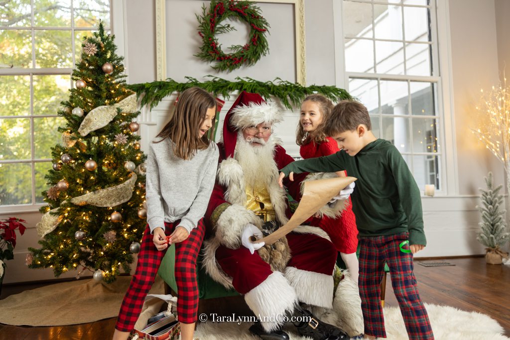 three kids gather around Santa to see if their name is on their list. Photo by Tara Lynn and Co. Photography